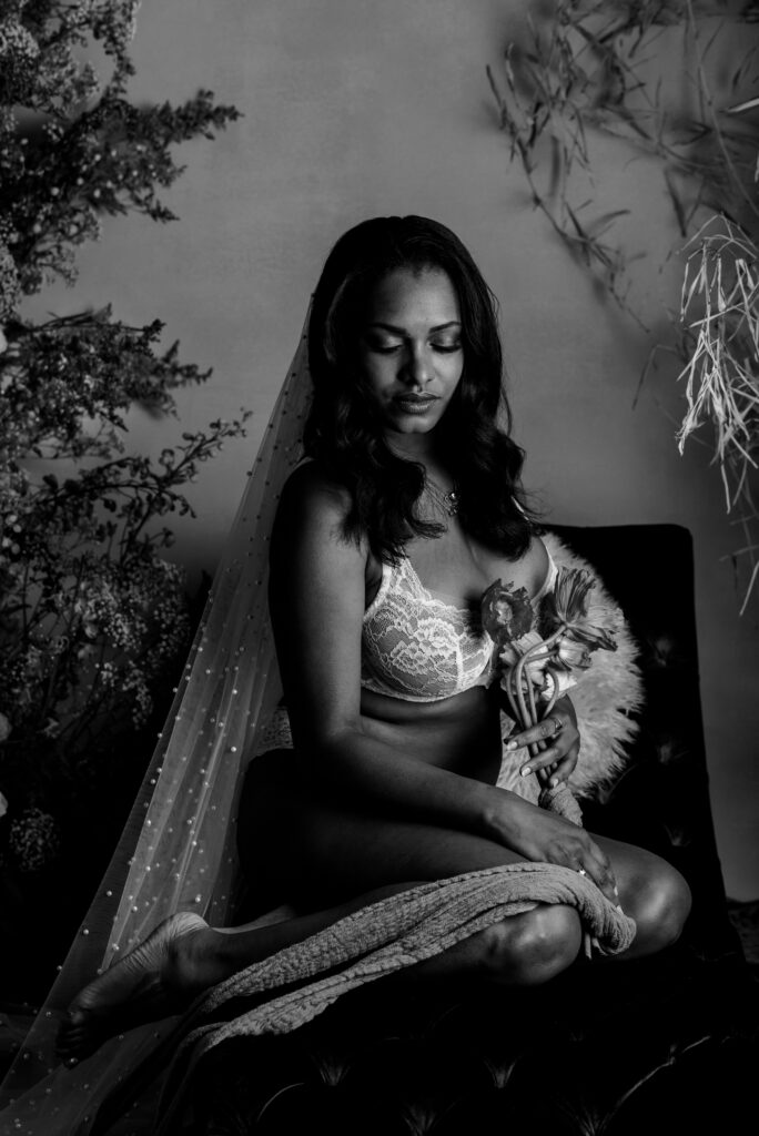 A woman in lacy bridal boudoir lingerie sits on a cushioned surface, draped in a sheer veil, holding a small bouquet. The black-and-white photo exudes a soft, elegant atmosphere with floral arrangements in the background.