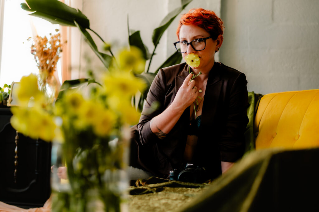Amy Rouyer of Wilde Company, a nashville boudoir photographer, A person with short red hair and glasses, wearing a dark jacket, holds a yellow flower to their lips while sitting on a yellow couch—an expressive moment of self-image captured through photography, with blurred yellow flowers in the foreground.