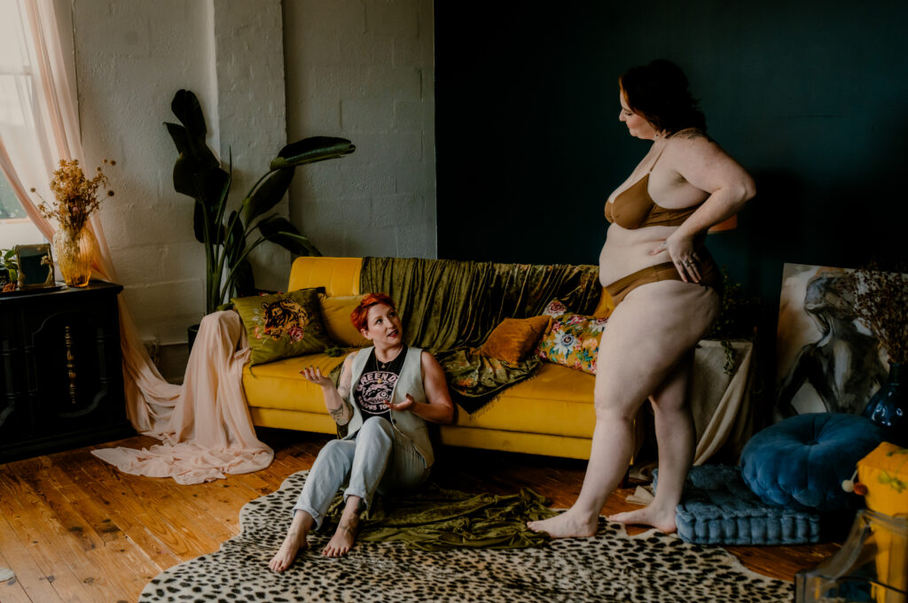 Two women interact in a cozy, sunlit room. One, standing in brown bridal boudoir underwear, faces the other, who sits on the floor by a yellow couch with colorful decor and plants; both appear engaged in conversation.