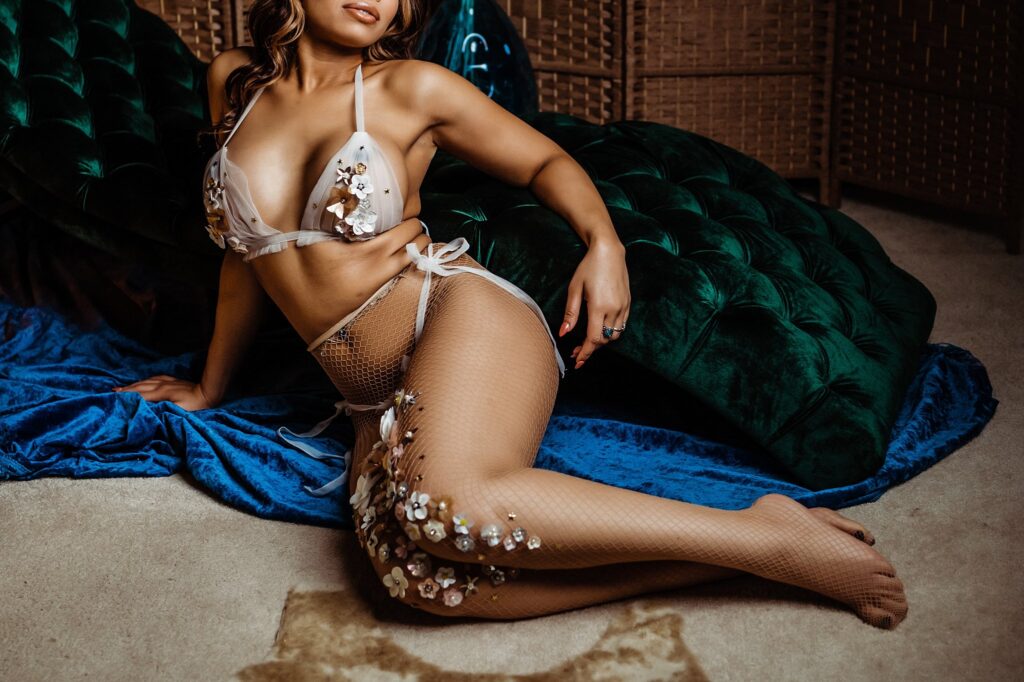 A woman poses on the floor in a sheer, floral-embellished lingerie set and fishnet tights—perfect for bridal boudoir—against a green velvet couch and blue fabric backdrop. Her face is partially out of frame.