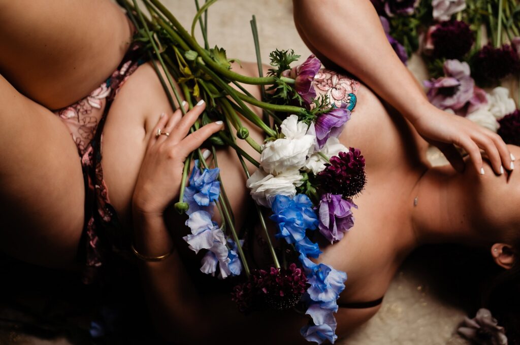 A person lying on their back, partially clothed in bridal boudoir lingerie, with colorful flowers—blue, white, and purple blooms—arranged across their torso and hand.
