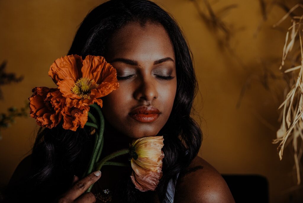 A woman with long dark hair gently holds two orange poppies near her face, eyes closed in a serene, bridal boudoir-inspired moment against a warm yellow-brown background.