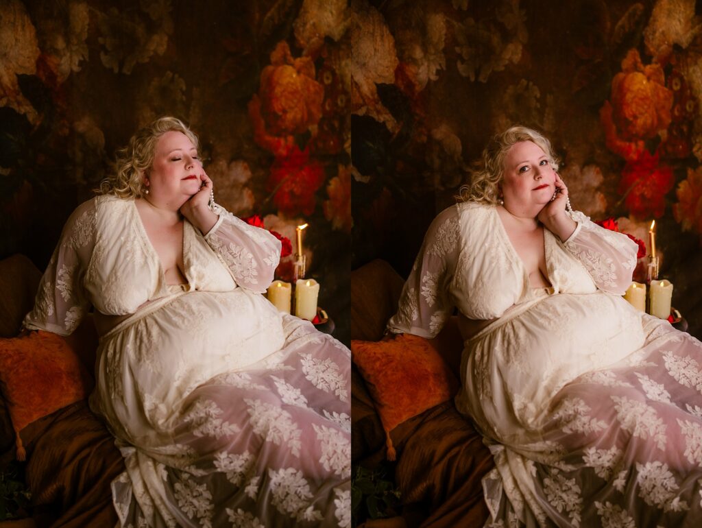 A woman in a sheer white lace dress sits on a bed surrounded by pillows and candles, resting her chin on her hand—an intimate bridal boudoir scene set against a dark, floral-patterned background.