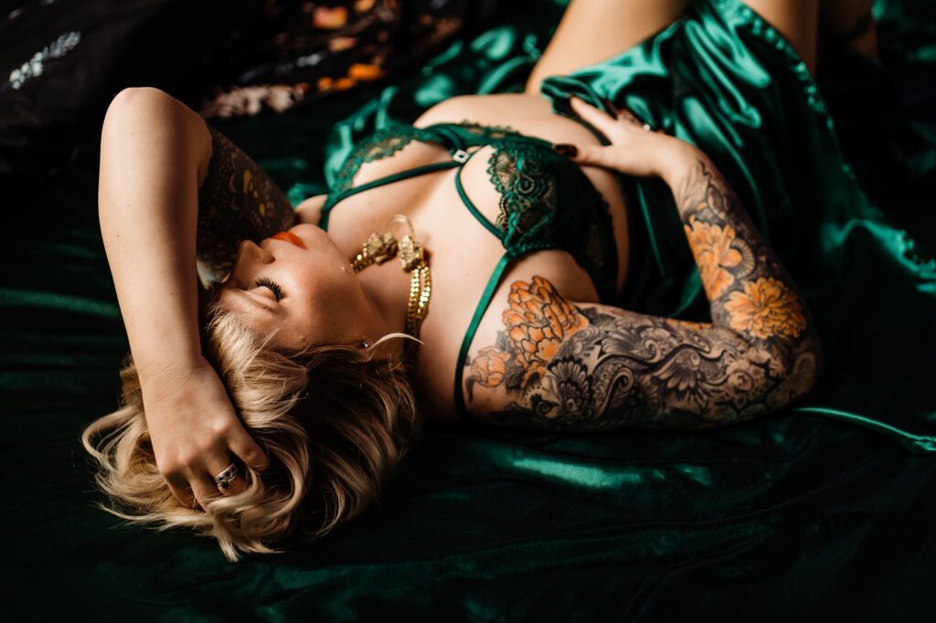 A person with blonde hair and tattooed arms lies on a dark green satin sheet, dressed in green lace lingerie, bridal boudoir style, with one hand on their head and the other resting on their body. Gold jewelry adorns their neck.