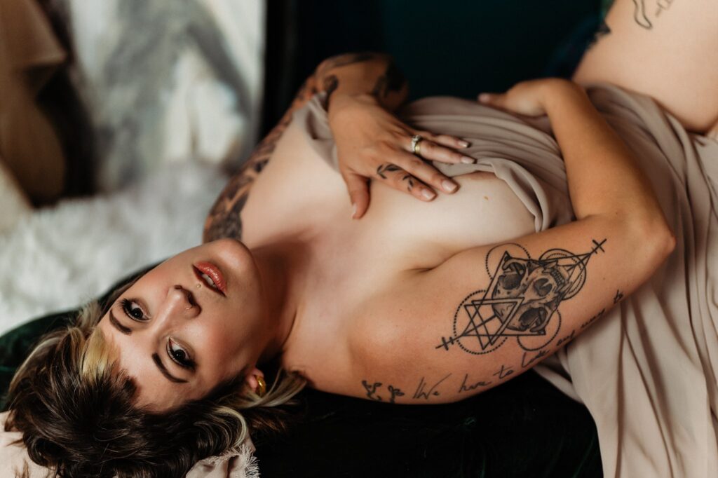 A person with tattoos lies on a green surface, partially covered by a beige fabric in a softly lit, intimate bridal boudoir setting. One hand covers their chest, while the other rests on their abdomen as they look at the camera.