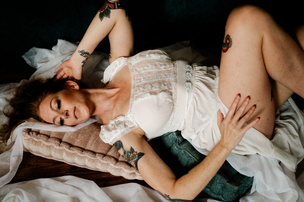 A woman with tattoos wearing a white lace dress lies on cushioned pillows, eyes closed, in a serene bridal boudoir setting, with one hand on her hip and the other by her head, surrounded by draped white fabric.