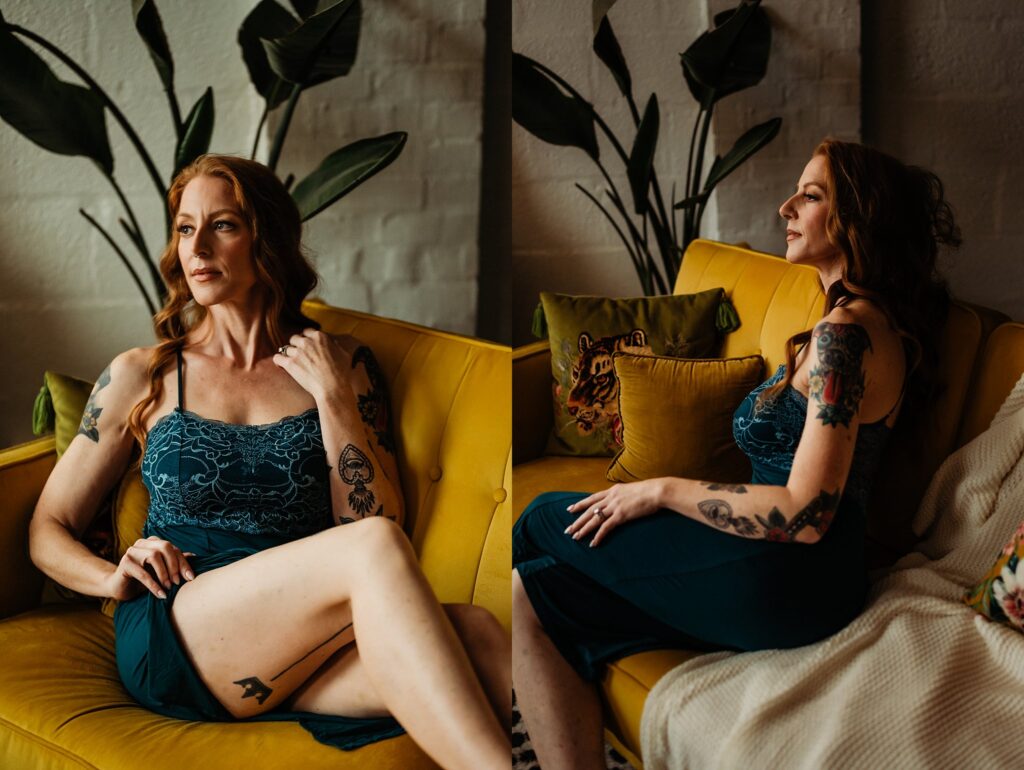 A woman with red hair and visible tattoos sits on a yellow sofa in a dark teal dress, exuding a bridal boudoir vibe. She gazes thoughtfully to the side, surrounded by green plants and pillows in the warmly lit, cozy setting.