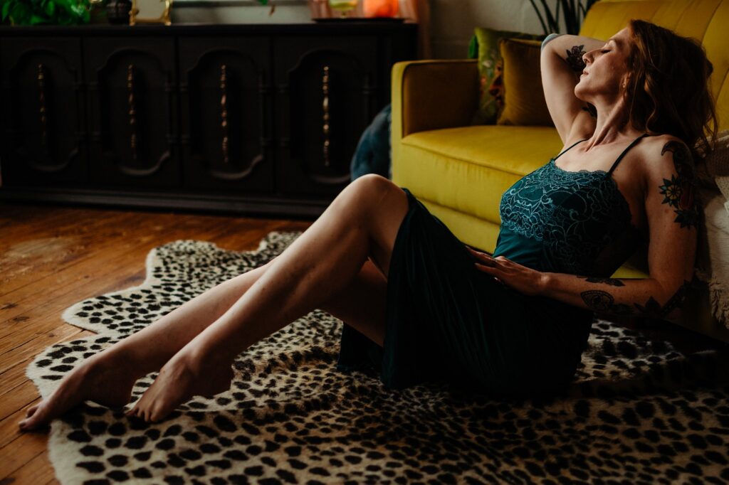 A woman in a dark dress sits on an animal-print rug, leaning back against a yellow couch with one arm raised and her eyes closed, capturing a quiet, intimate moment in a dimly lit bridal boudoir with wooden floors and decorative furniture.
