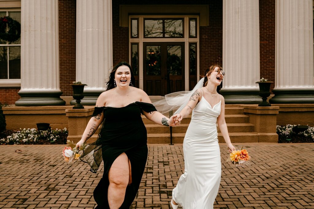 Two women, one in a black dress and one in a white wedding gown with a veil, hold hands and smile joyfully while walking outside a building with large columns—captured beautifully by a talented Nashville wedding photographer.