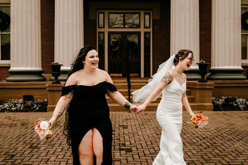 Two joyful brides, captured by a Nashville wedding photographer, laugh and hold hands while walking outside a building with large columns—one in a white dress and veil, the other in a black off-the-shoulder gown—each holding a bouquet.