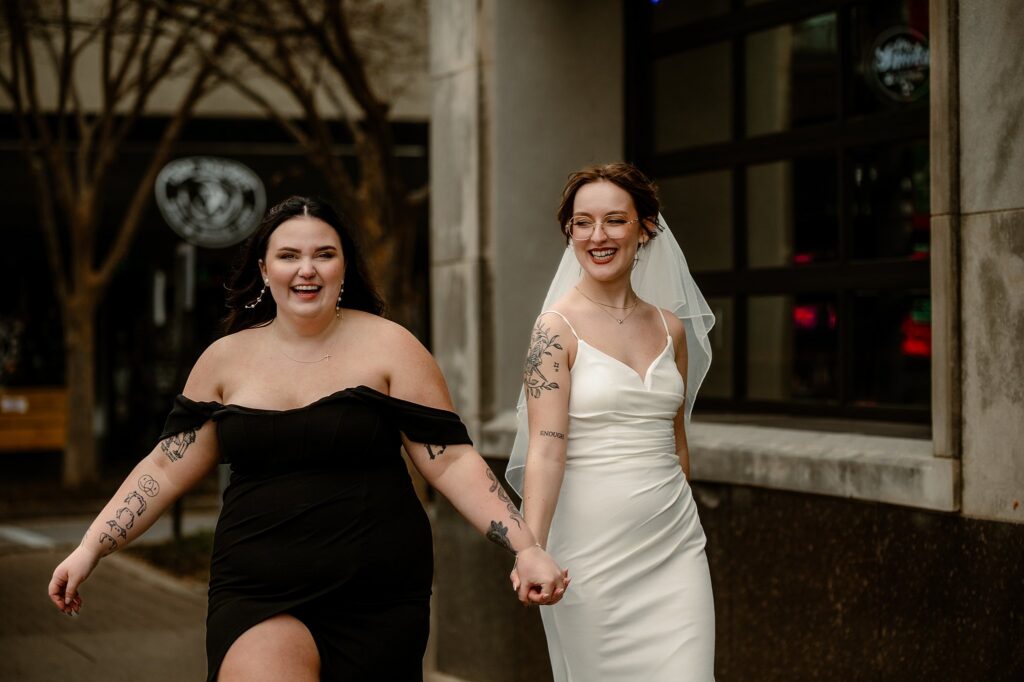 Two women are smiling and holding hands while walking outside. Captured by a Nashville wedding photographer, one wears an off-shoulder black dress and the other a white wedding dress with a veil. Both have visible tattoos and look happy.