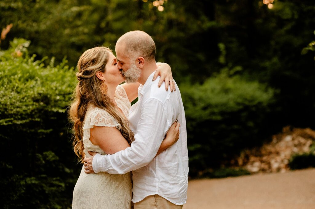 A couple embraces and kisses outdoors as a Nashville wedding photographer captures the moment. The woman wears a cream lace dress, and the man wears a white shirt and beige pants. They stand closely together, surrounded by lush greenery.