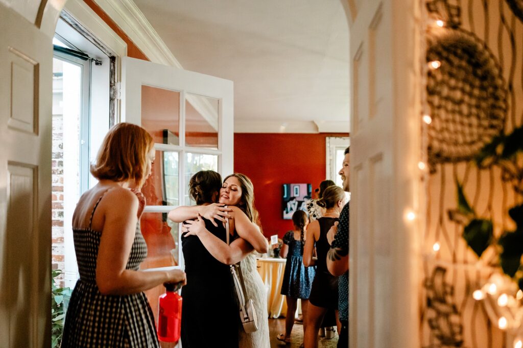 A group of women gather indoors at a brightly lit social event. Two women hug near the doorway while others chat in the background, captured perfectly by a Nashville wedding photographer. String lights and decor add a warm, festive atmosphere.
