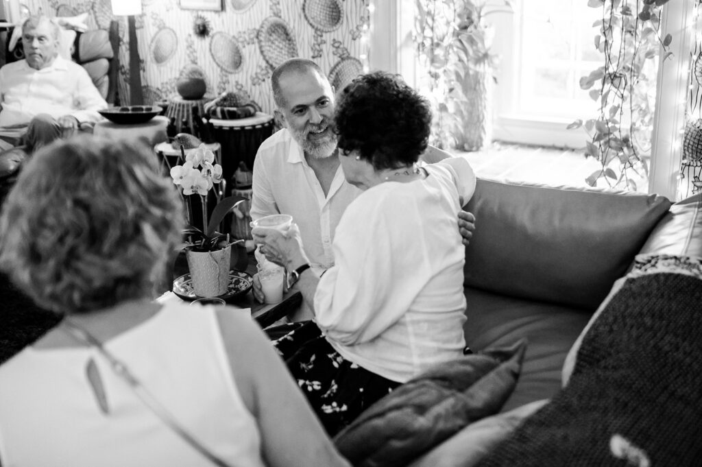 A black-and-white photo by a Nashville wedding photographer shows three people on a couch, deep in conversation. A man faces two women; one holds a drink. In the background, a fourth person sits in the cozy, plant-filled living room.