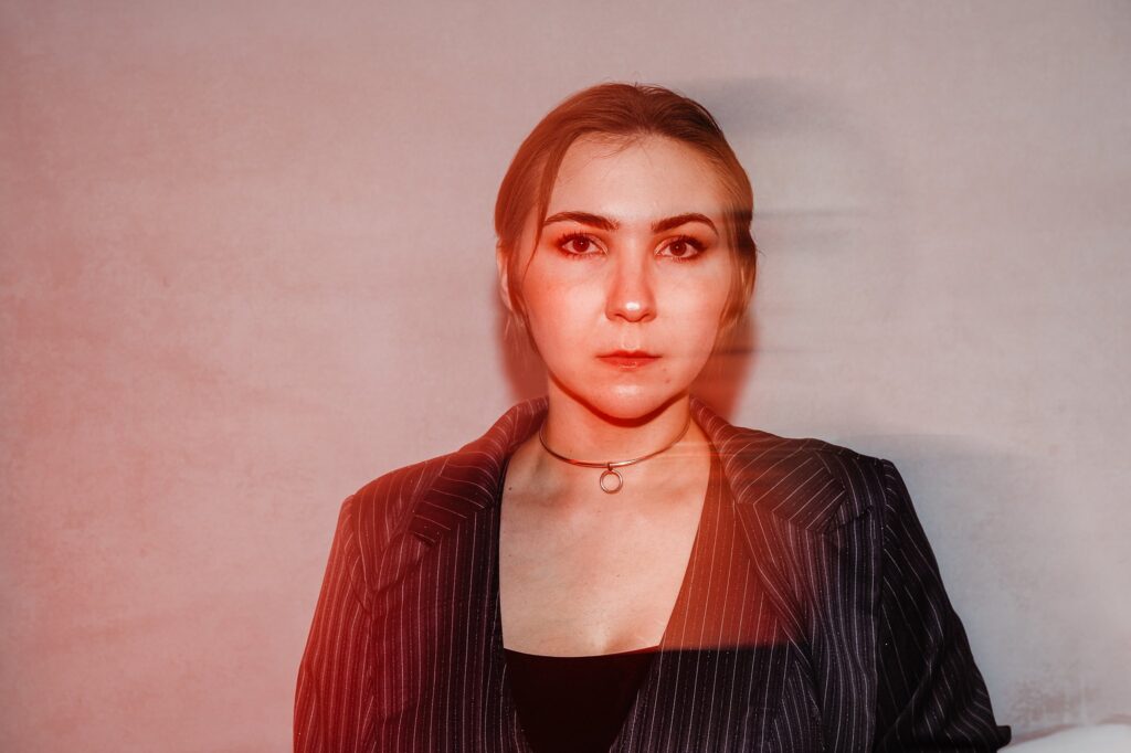A woman with light skin and blonde hair wears a pinstripe blazer and choker necklace, standing against a plain light background. The image, by a Nashville creative portrait photographer, features a red tint and a blurred effect around her face.