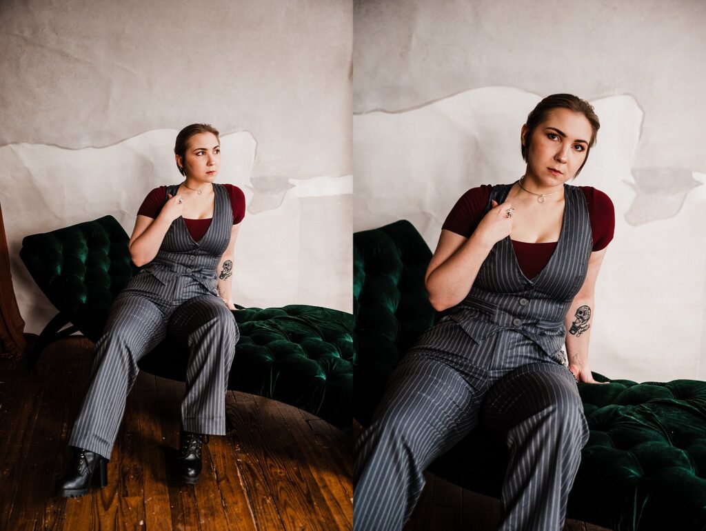 A woman with light brown hair in a low bun sits on a dark green velvet couch. Photographed by a Nashville creative portrait photographer, she wears a pinstriped suit over a burgundy shirt and black boots, tattoos visible on her left arm.