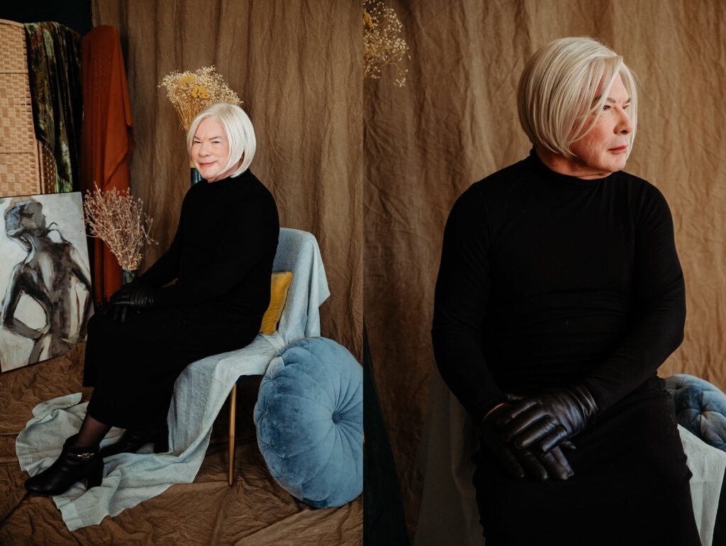 A person with short white hair in a black dress, gloves, and shoes sits on a chair with colorful fabrics and pillows, posing against a brown draped backdrop with dried flowers and a painting—styled by a Nashville creative portrait photographer.