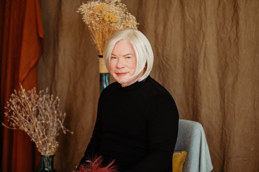 An older person with light skin and straight, white hair sits on a chair in a black turtleneck. Dried flowers and brown drapes set a warm, cozy mood—perfectly captured by a Nashville creative portrait photographer.