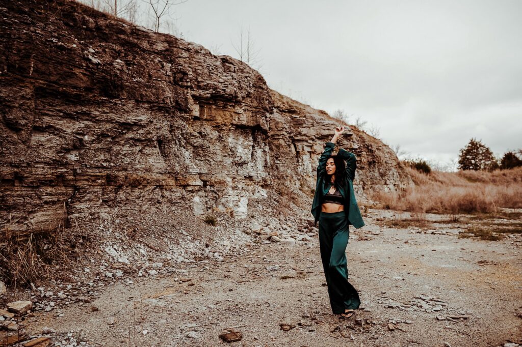A woman in a green outfit stands with one arm raised in an outdoor rocky area, cliffs and dry grass behind her beneath a cloudy sky, captured by a Nashville creative portrait photographer.
