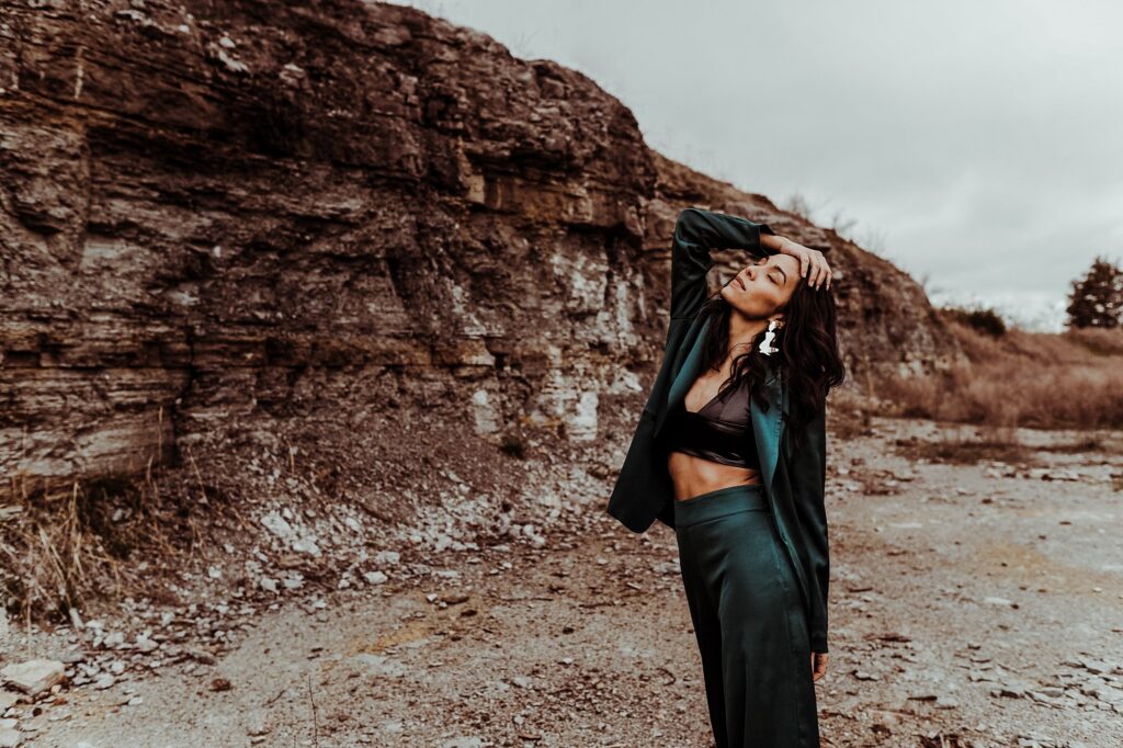 A woman in a dark green outfit poses outdoors before a rocky cliff, standing on gravel with one hand on her head and eyes closed beneath a cloudy sky, captured by a Nashville creative portrait photographer.