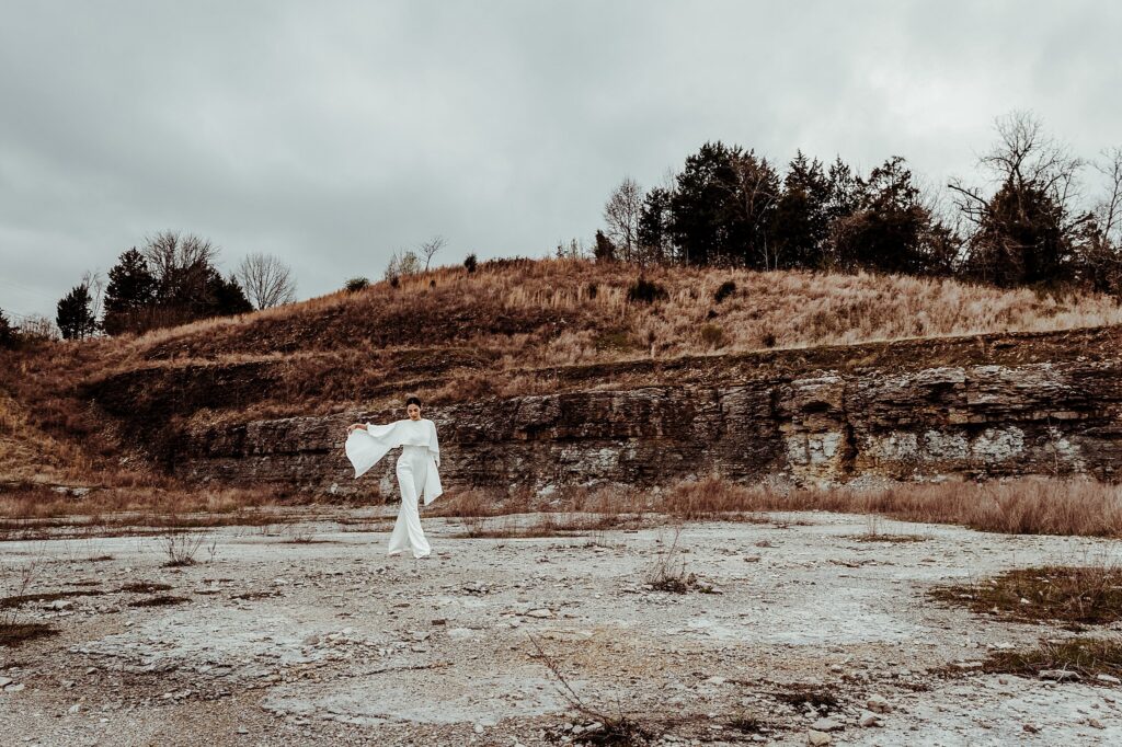 A person dressed in flowing white clothing stands in the center of a barren, rocky landscape with grassy hills and sparse trees under a cloudy sky—captured by a Nashville creative portrait photographer.