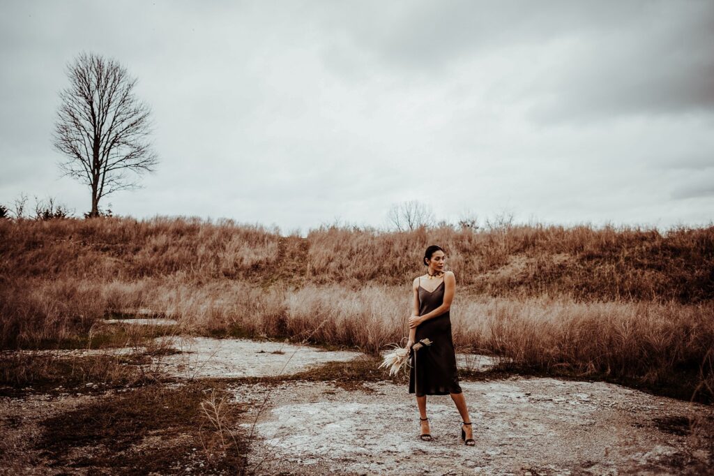A woman in a dark dress stands alone in a barren, grassy field, holding a bouquet of dried flowers. The overcast sky and lone tree create a moody scene, captured by a Nashville creative portrait photographer.