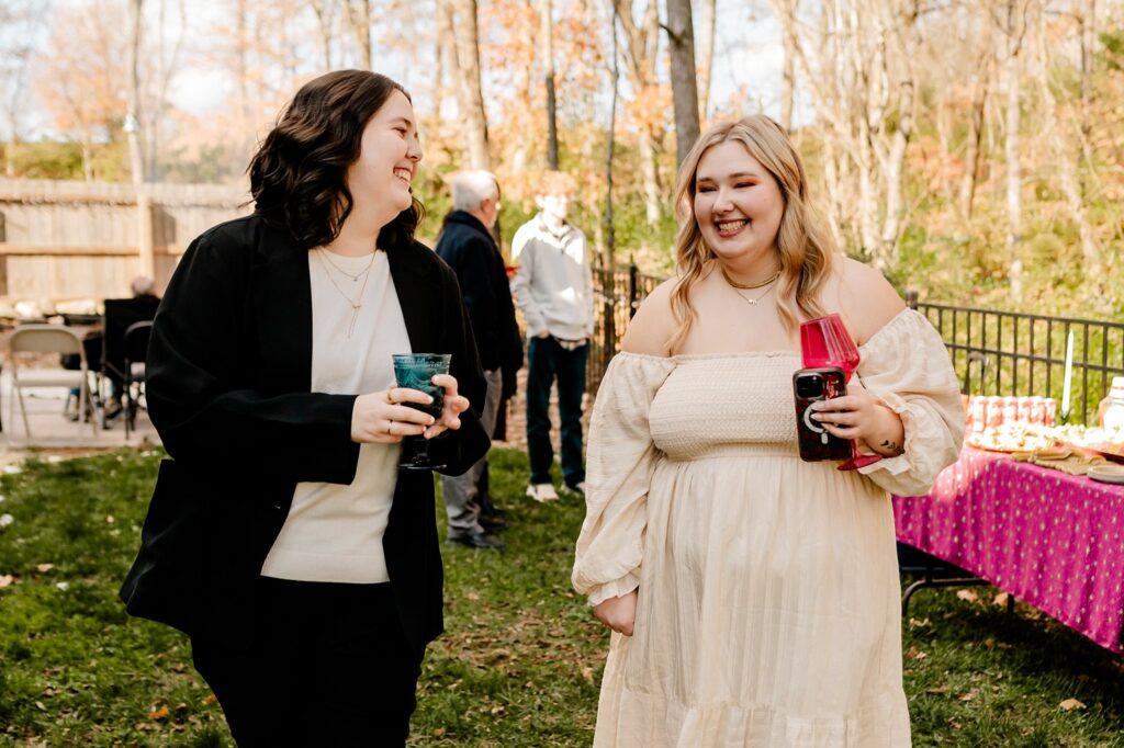 Two people are smiling and walking outdoors at a gathering, captured by a Nashville wedding photographer. One wears a black suit, the other a white off-shoulder dress. Both hold drinks as guests mingle near a pink-covered table with snacks in the background.