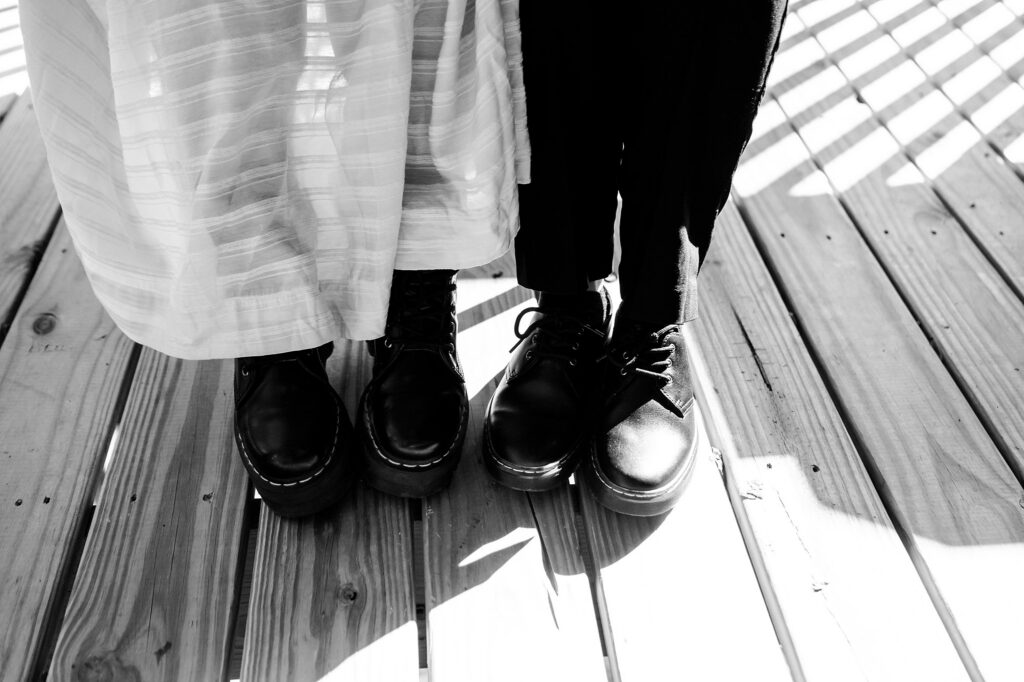 Two people stand close together on a wooden deck, sunlight casting striped shadows. One wears a long, light-colored skirt; the other, dark pants—both in black shoes. Captured with the artistry of a Nashville wedding photographer.