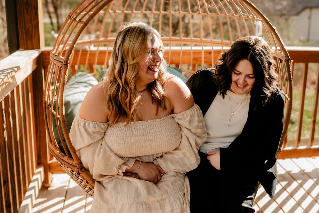 Two women sit together on a hanging wicker chair outdoors, laughing in the sunlight. Captured by a Nashville wedding photographer, one wears an off-shoulder beige dress, the other a black jacket over white. The background shows a wooden deck.