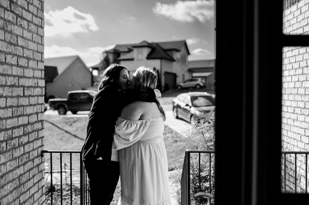 Two people embrace on a porch in front of a house, one in a dark suit and the other in a light off-shoulder dress. Captured by a Nashville wedding photographer, the daytime scene includes cars and houses in the background.
