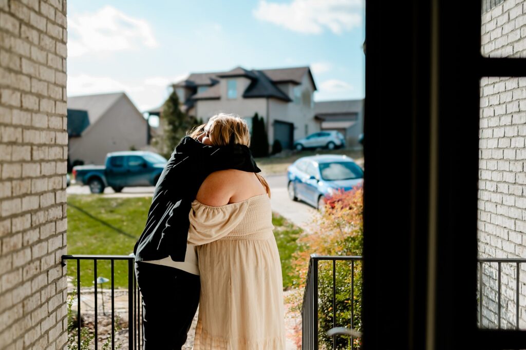 Two people embrace on a front porch, framed by brick walls, as suburban houses and cars sit beneath a bright sky—captured in the warm, candid style of a Nashville wedding photographer.
