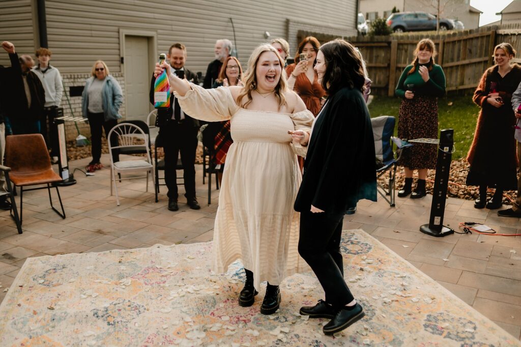 Two people dance joyfully on a rug outdoors as friends smile and clap. One, in a cream dress, waves a colorful flag; the other wears dark clothing. Captured beautifully by a Nashville wedding photographer, it’s a backyard celebration to remember.