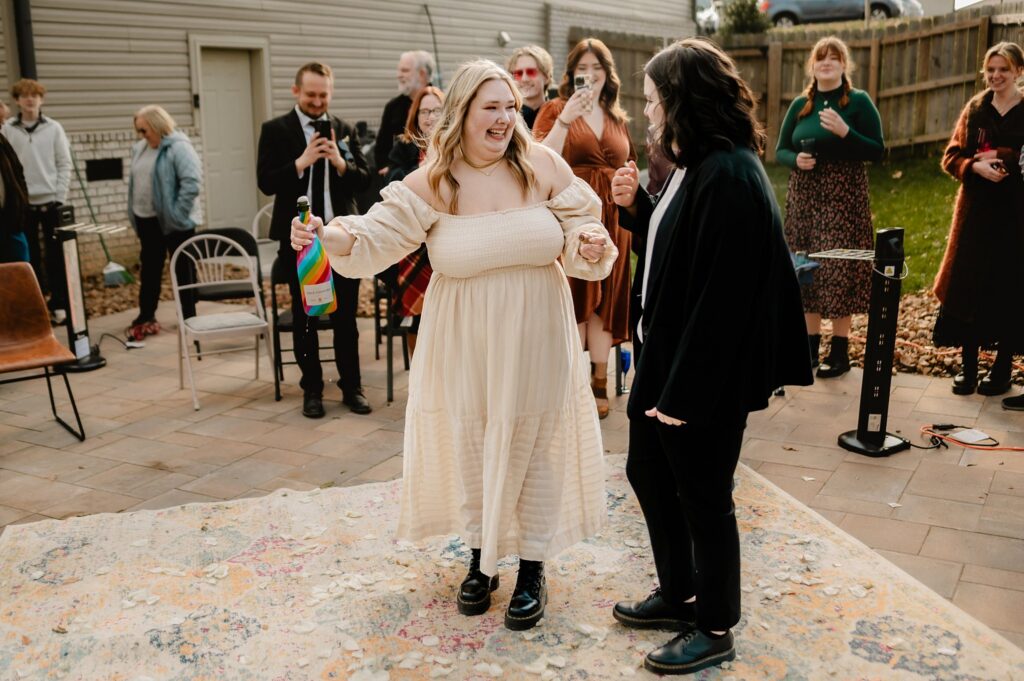 Two people, one in a cream dress and one in black pants and a jacket, smile and hold hands on a rug outdoors. The person in the cream dress holds a bottle as others watch and take photos, captured by a Nashville wedding photographer.
