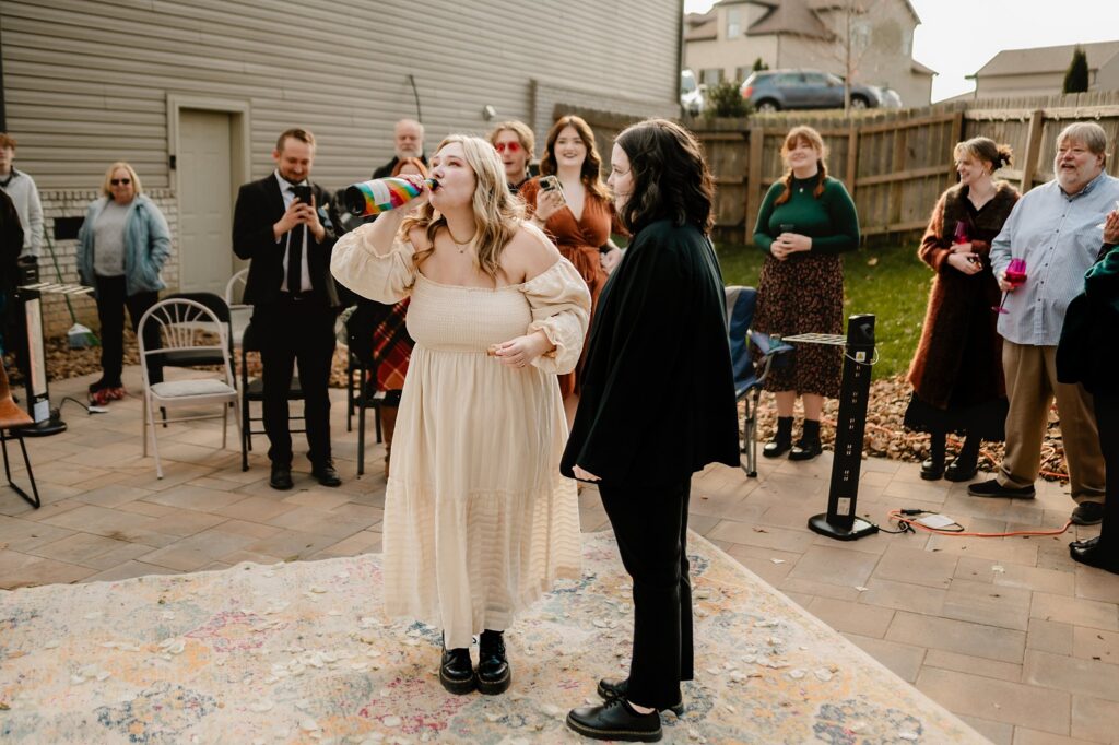 A woman in a cream dress drinks from a colorful bottle while people stand around her outdoors, smiling and watching. Captured by a Nashville wedding photographer, the group is gathered on a patio with houses and a wooden fence in the background.