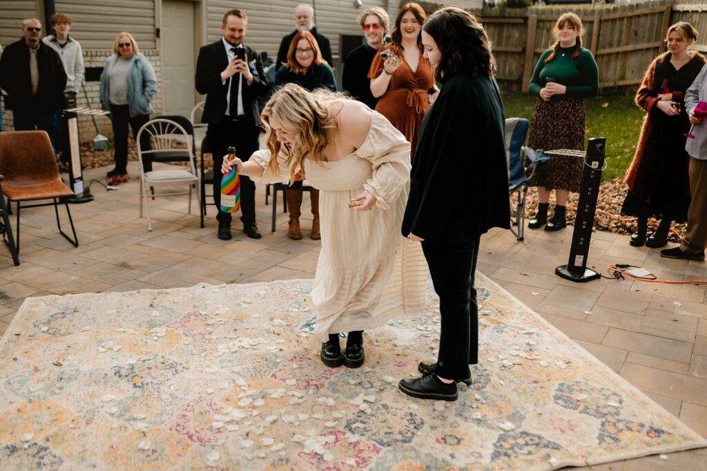 A person in a cream dress bends down to pick up something from a rug while another stands nearby; a group of people watch them outdoors, smiling in cool weather—captured beautifully by a Nashville wedding photographer.