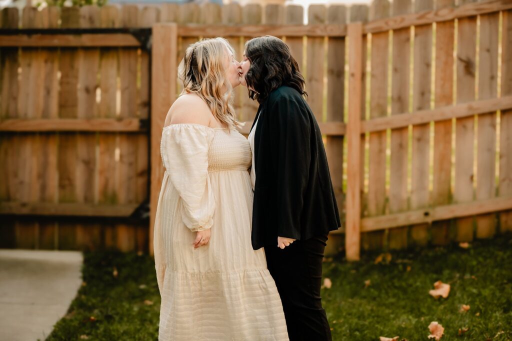 Two women share a warm, intimate kiss in a backyard with a wooden fence—one in a white off-the-shoulder dress, the other in a black jacket and pants. Captured beautifully by a Nashville wedding photographer.
