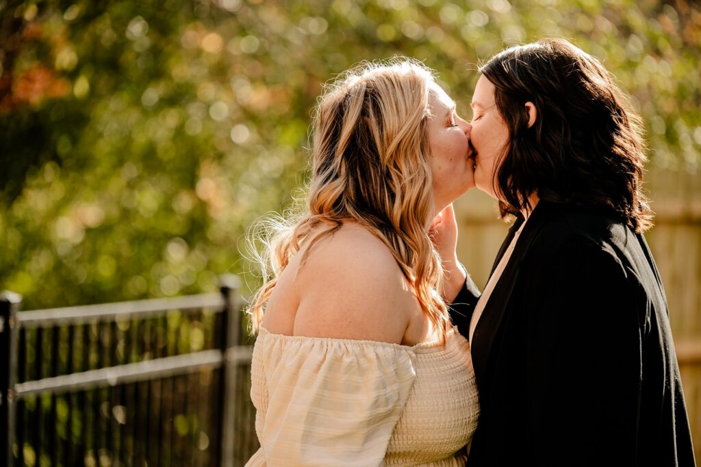 Two women share a kiss outdoors in warm sunlight, standing close together by a fence with green foliage in the background—a beautiful moment captured by a Nashville wedding photographer. One wears an off-the-shoulder dress, the other a dark jacket.