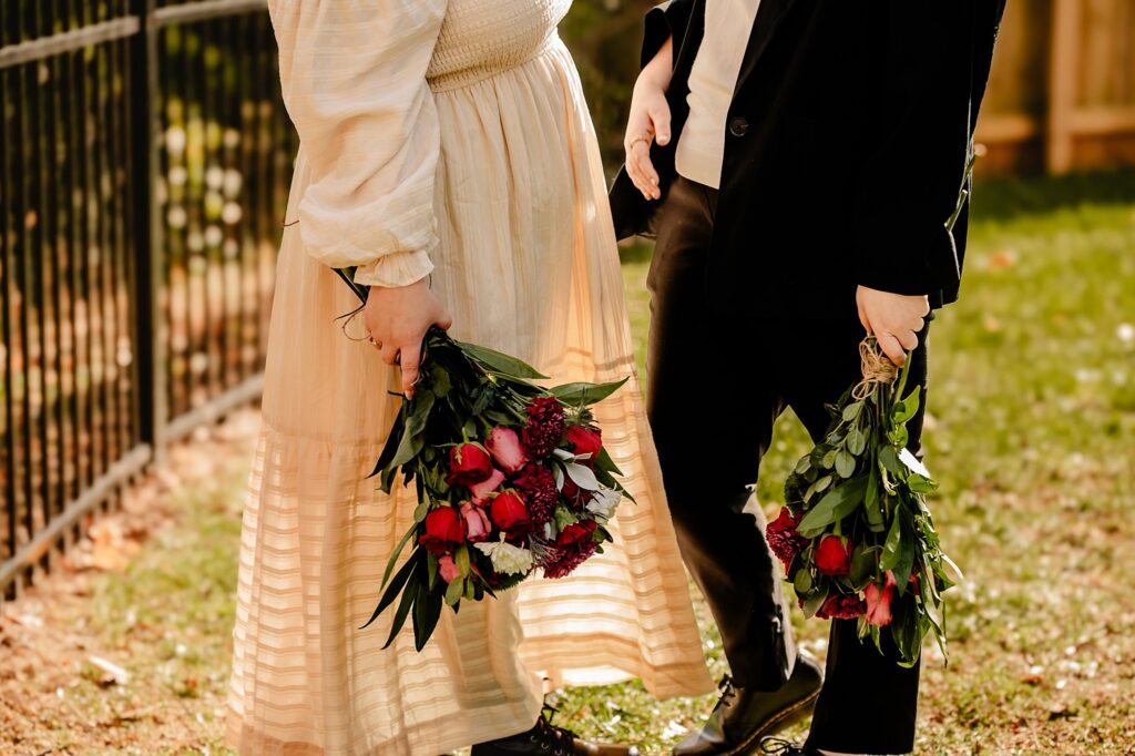 Two people stand outdoors, vibrant flower bouquets in hand—one in a cream dress, the other in a black suit. Sunlight illuminates their moment, perfectly captured by a Nashville wedding photographer.