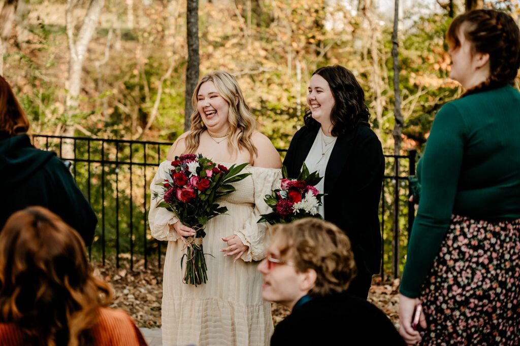 Two women stand outdoors, smiling and holding bouquets of flowers, captured by a Nashville wedding photographer. Surrounded by guests, autumn trees, and a black metal fence, they appear to be at a joyful celebration or gathering.