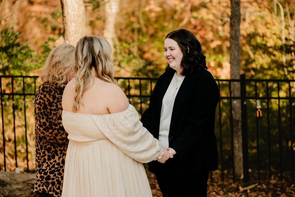 Two women hold hands and smile at each other during a ceremony, as a person in animal print officiates. Captured by a Nashville wedding photographer, the moment unfolds outdoors amid autumn trees and a black metal fence.