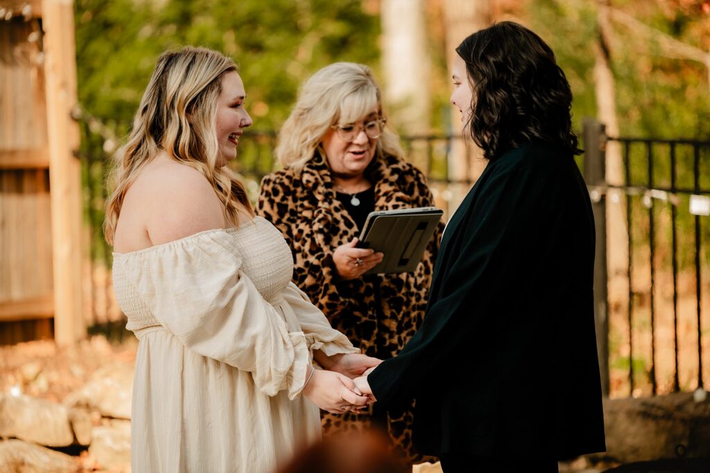 Two people stand facing each other, holding hands and smiling during an outdoor ceremony. A nashville wedding photographer captures the moment as an officiant in a leopard print coat reads from a tablet with trees and a fence behind them.