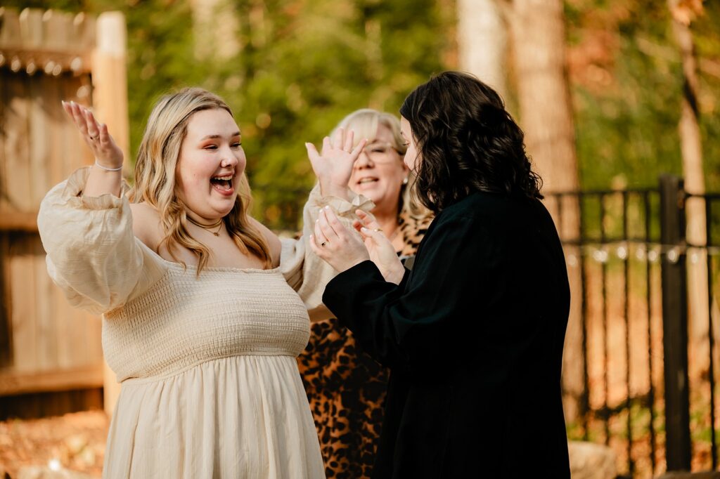 Two brides stand outdoors, smiling and raising their hands in excitement. Captured by a Nashville wedding photographer, one wears a cream dress, another a leopard print top, and the third a dark jacket. Trees and a fence are visible in the background.