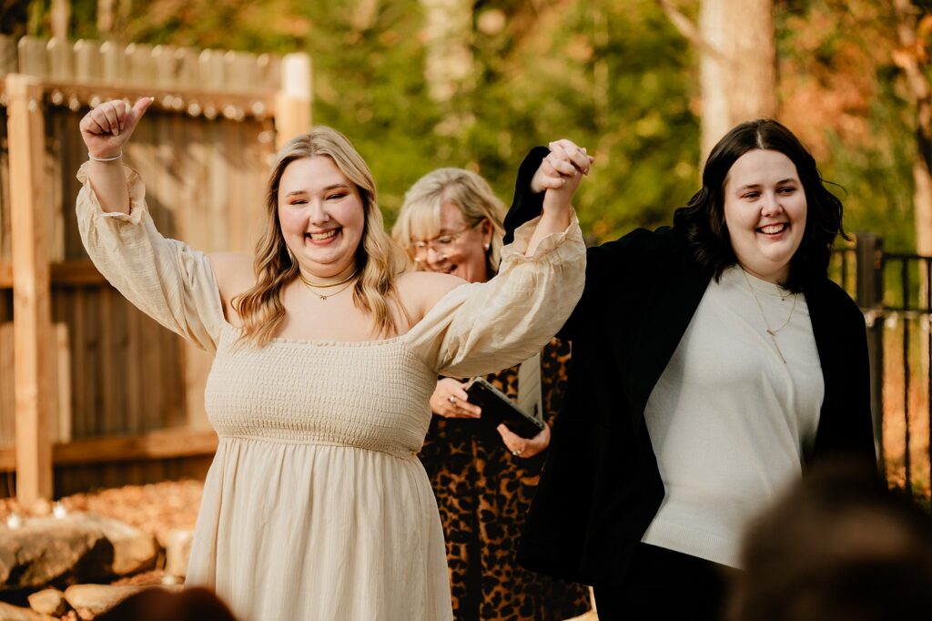 Two smiling brides hold hands and raise their arms in celebration outdoors, captured perfectly by a Nashville wedding photographer, with a woman in glasses and animal print standing behind them. Trees and a wooden fence are visible in the background.