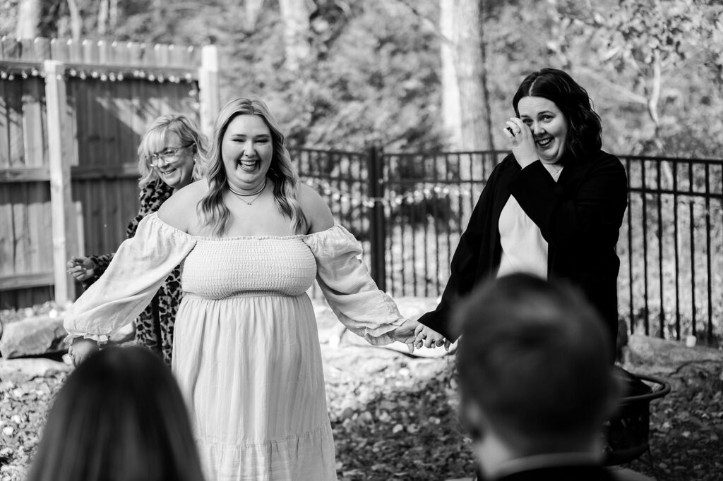 Two brides, one in a white off-shoulder dress, smile and hold hands outdoors near a fence. As another woman wipes a tear, blurred guests look on—capturing this joyful, emotional moment as only a skilled Nashville wedding photographer can.