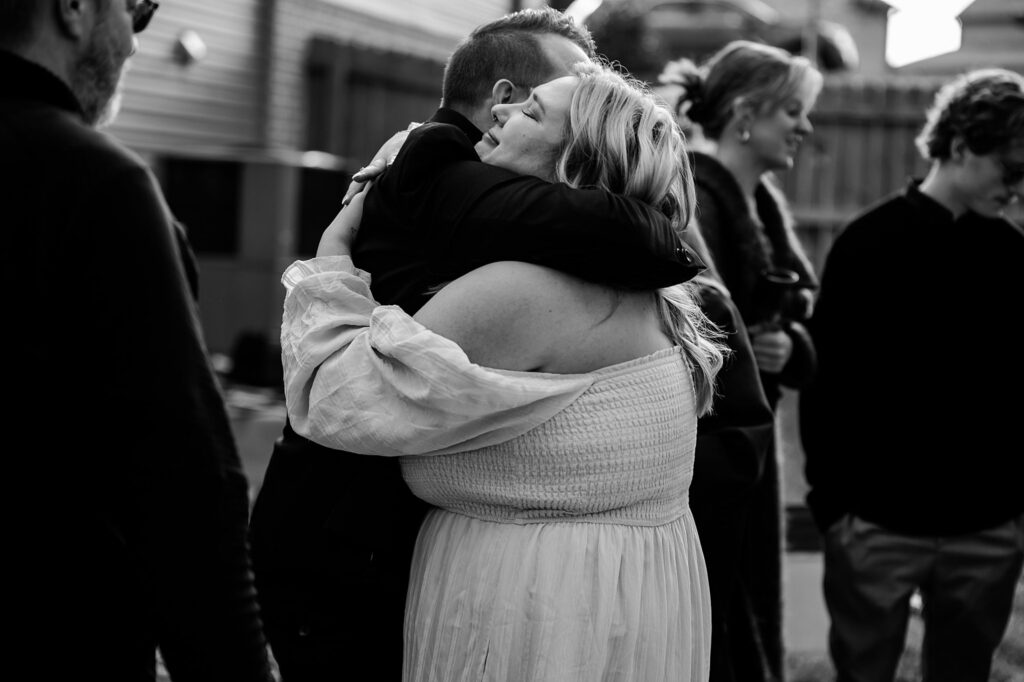 A bride in a light-colored dress warmly hugs someone in a dark jacket at an outdoor gathering, captured by a Nashville wedding photographer. Others stand and talk in the black-and-white scene.