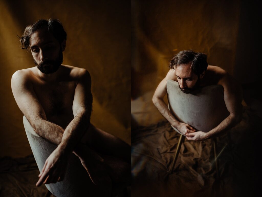 A man with brown hair and a beard sits nude behind a light-colored chair, his arms and body partially obscured. Dimly lit with a warm, brown backdrop, this image captures the artistic style of a Nashville creative portrait photographer.