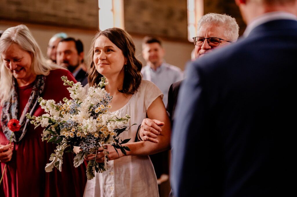 A bride holding a bouquet walks down the aisle arm in arm with an older man, smiling, as guests look on in a warmly lit indoor venue—beautifully captured by a Nashville wedding photographer.