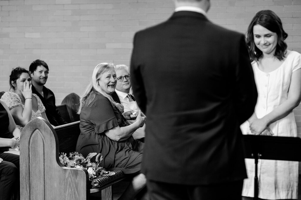A group of people sit in pews, smiling and laughing, during what appears to be a wedding or formal ceremony. Captured by a Nashville wedding photographer, the joyful moment shines as a woman in the front row smiles at the couple. The image is in black and white.