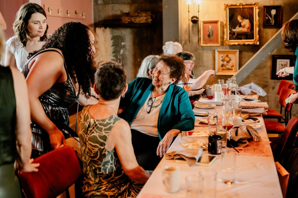 A group of women gathered around a smiling elderly woman at a long table, engaging in conversation in a warmly lit room adorned with paintings—captured beautifully by a Nashville wedding photographer.