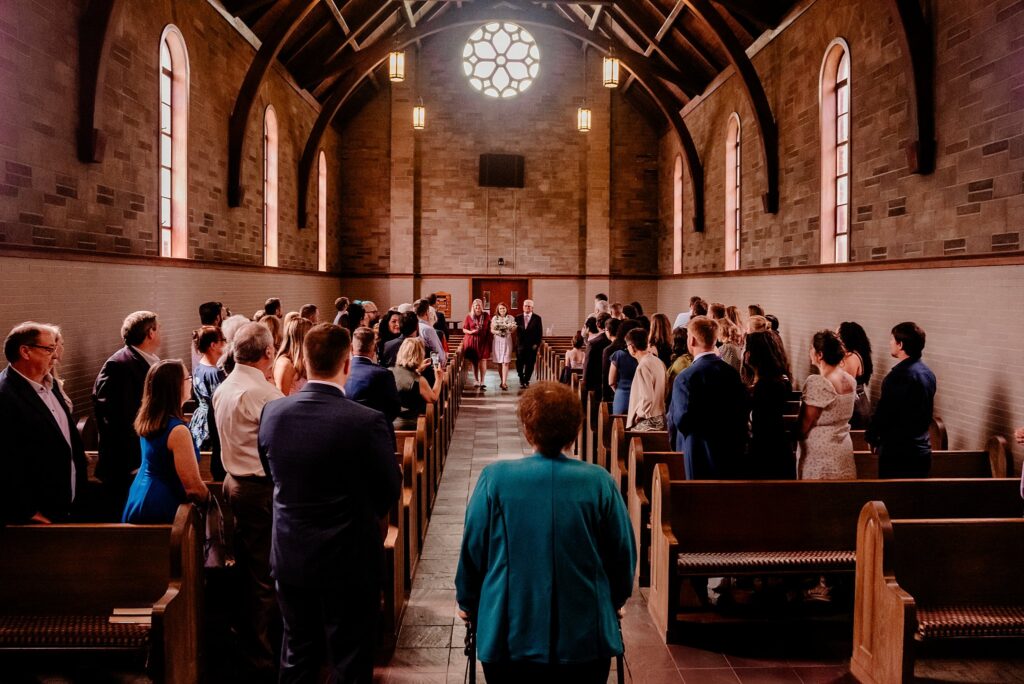 Guests stand in pews inside a church with arched ceilings and stained glass windows as a Nashville wedding photographer captures the ceremony unfolding at the front altar.